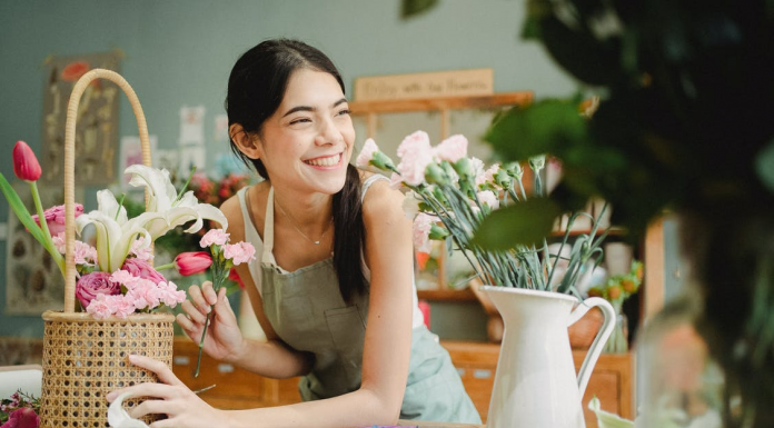 a smiling lady is sitted on the table where beautiful flowers are keeping in the basket to hangout in the home decor enhancement